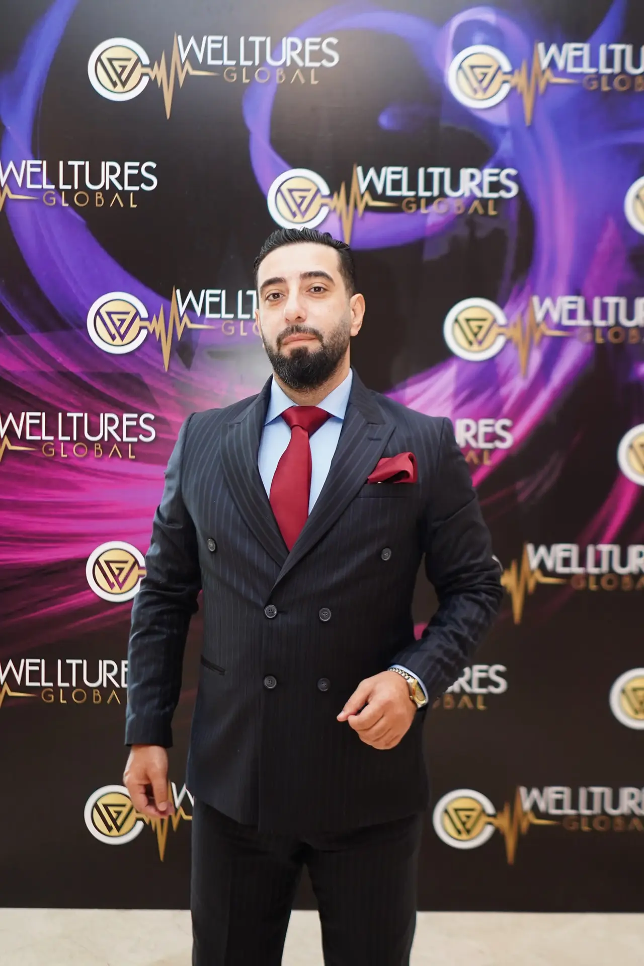 Welltures Global basın duvarı önünde lacivert çizgili takım ve kırmızı kravatla